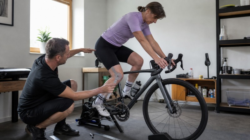 Cyklista sedí na kole v hale a bikefitter mu měří úhel kolena při šlapání